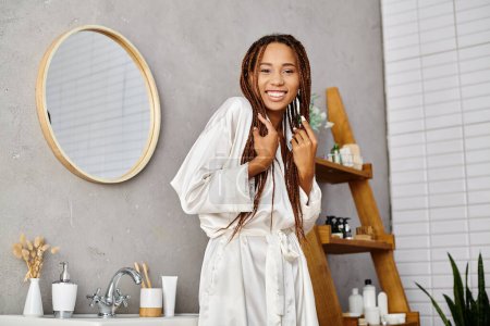 African American woman with afro braids standing in front of modern bathroom sink in bath robe, focusing on beauty and hygiene.
