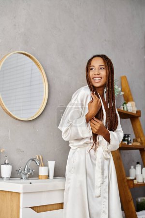 Afro-Amerikanerin mit Afro-Zöpfen im Bademantel vor modernem Badezimmerwaschbecken und Fokus auf Schönheit und Hygiene.