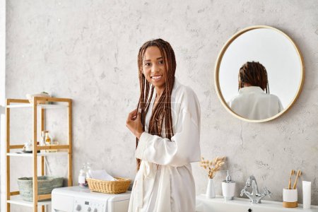 An African American woman with Afro braids stands in front of a mirror in a modern bathroom, wearing a bathrobe.