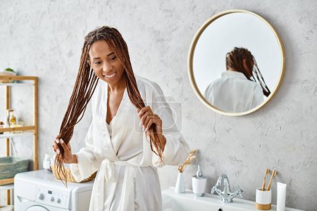 Afroamerykanka w afro warkoczach w szlafroku, stoi przed nowoczesnym umywalką łazienkową, podziwiając jej odbicie.