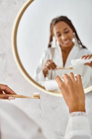 An African American woman with afro braids brushes her teeth in front of a mirror in a modern bathroom.