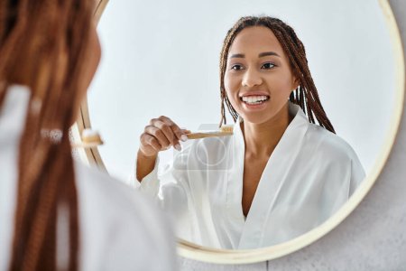 Afroamerykanka z afro warkoczami, w szlafroku, myjąca zęby w nowoczesnym lustrze w łazience.