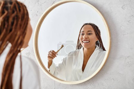 An African American woman with afro braids in a bathrobe brushing her teeth in front of a mirror in a modern bathroom.