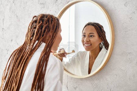 An African American woman with afro braids in a bath robe brushing her teeth in a modern bathroom mirror.