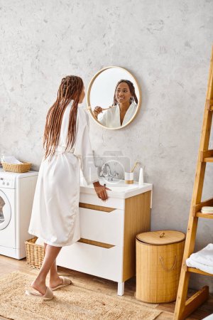 An African American woman stands in her modern bathroom, admiring herself in the mirror while wearing a bathrobe.