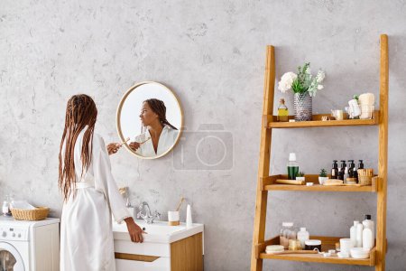 Une femme afro-américaine élégante dans un peignoir avec des tresses afro debout devant un lavabo de salle de bains moderne.