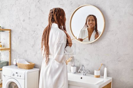 Téléchargez les photos : Une Afro-Américaine avec des tresses afro se tient devant un miroir dans une salle de bain moderne tout en portant un peignoir. - en image libre de droit