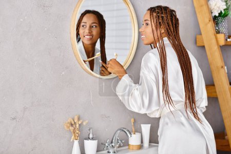 An African American woman with afro braids in a bathrobe brushing her teeth in a modern bathroom in front of a mirror.