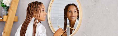 Téléchargez les photos : Femme afro-américaine avec des tresses afro brosse ses cheveux dans une salle de bain moderne portant un peignoir. - en image libre de droit
