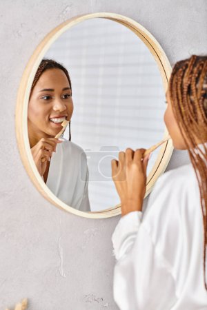 Eine afroamerikanische Frau mit Afro-Zöpfen putzt sich in einem modernen Badezimmer die Zähne, trägt einen Bademantel.