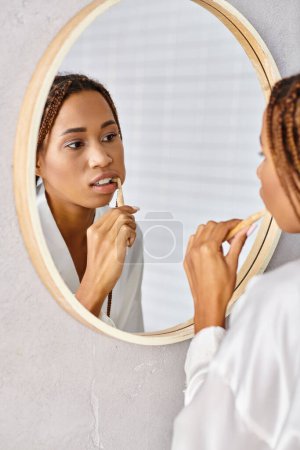 An African American woman with afro braids in a bathrobe brushing her teeth in a modern bathroom mirror.