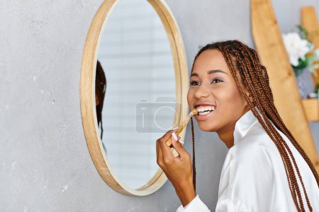 Una mujer afroamericana con trenzas afro en un albornoz cepillándose los dientes frente a un espejo en un baño moderno.