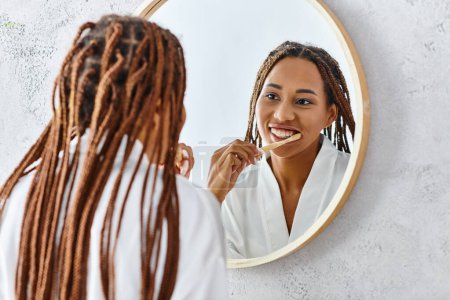 African American woman with afro braids in bath robe brushing teeth in front of a mirror in modern bathroom.