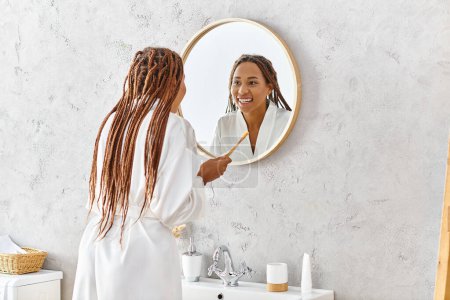 Femme afro-américaine avec tresses en peignoir de bain brossant les dents dans le miroir de salle de bain moderne.