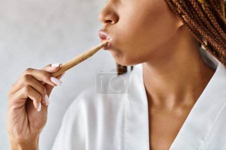 An African American woman with afro braids brushes her teeth with a wooden toothbrush in a modern bathroom wearing a bath robe.