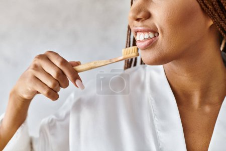 An African American woman with afro braids, in a bathrobe, brushing her teeth with a wooden toothbrush in a modern bathroom.