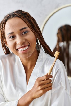 Une femme avec des dreadlocks dans un peignoir tient une brosse à dents dans une salle de bain moderne, mettant l'accent sur la beauté et l'hygiène.
