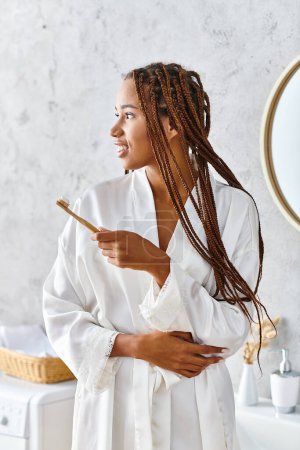 An African American woman with dreadlocks, dressed in a bathrobe, brushing her teeth in her modern bathroom.