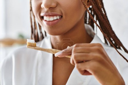 An African American woman in a bathrobe with afro braids brushes her teeth in a modern bathroom for beauty and hygiene.