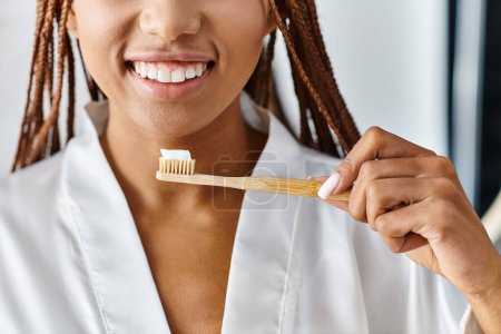 An African American woman with afro braids in a bathrobe brushing her teeth with a wooden toothbrush in a modern bathroom.