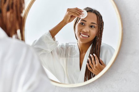 An African American woman with afro braids using cotton pad with toner in front of a mirror in a modern bathroom