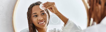 Photo for An African American woman with afro braids using cotton pad with toner in front of a mirror in a modern bathroom, focusing on beauty and hygiene. - Royalty Free Image