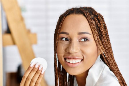 Téléchargez les photos : Femme afro-américaine avec des tresses en peignoir, tenant un objet blanc dans sa main dans une salle de bain moderne. - en image libre de droit