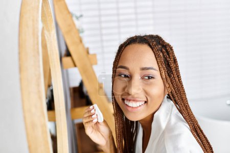 An African American woman with afro braids smiles holding a toothbrush in a modern bathroom, exuding joy and hygiene.