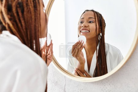 Una mujer afroamericana con trenzas afro en un albornoz con almohadilla de algodón frente a un espejo en un baño moderno.