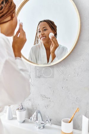 Foto de Una mujer afroamericana con trenzas afro usando almohadilla de algodón frente a un espejo en un baño moderno. - Imagen libre de derechos