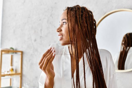 African American woman with afro braids stands in front of mirror in bathrobe, holding cotton pad in modern bathroom.