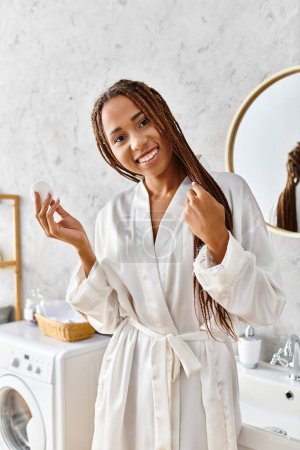 Una mujer afroamericana con trenzas afro en un albornoz sosteniendo pap de algodón en un baño moderno, centrándose en la belleza y la higiene.