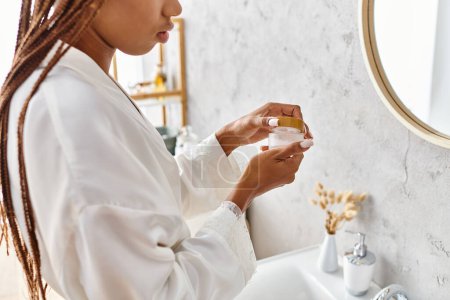 Photo for African American woman with afro braids in white robe holding beauty jar with cream in modern bathroom for beauty and hygiene. - Royalty Free Image