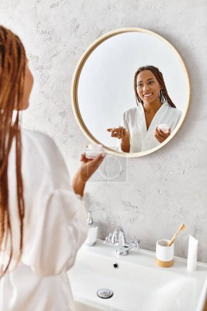 Eine afroamerikanische Frau mit Afro-Zöpfen steht in einem modernen Badezimmer und hält ein Schönheitsglas mit Creme in der Hand