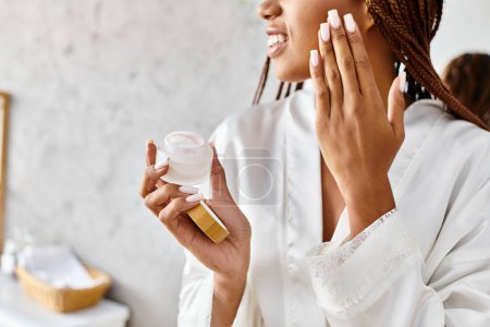African American woman with afro braids in bathrobe holding a jar of cream in front of her face in a modern bathroom.