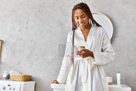African American woman in bathrobe holding beauty jar with cream in modern bathroom.