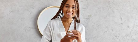 A stylish woman with dreadlocks holds a glass jar in front of a mirror in a modern bathroom, exuding beauty and confidence.