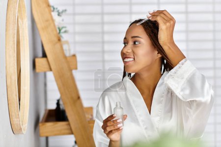 An African American woman with afro braids is holding a bottle with serum in her modern bathroom while wearing a bath robe.