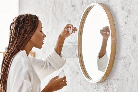 An African American woman with Afro braids in a bathrobe applying serum in a modern bathroom.
