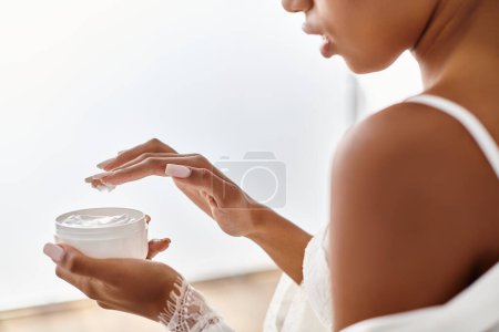 An African American woman with afro braids peacefully holds a cream jar in her hands in a modern bathroom.