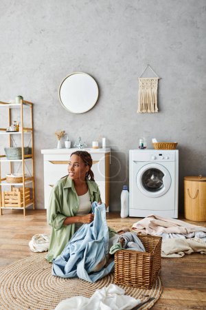 An African American woman with afro braids sitting on the floor in front of a washing machine, doing laundry in a bathroom.