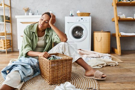 Une Afro-Américaine avec des tresses afro est assise à côté d'un panier à linge dans la salle de bain, se préparant à laver les vêtements.