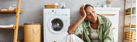 Foto de Una mujer afroamericana con trenzas afro se sienta en el suelo, con una lavadora en el fondo, haciendo tareas domésticas. - Imagen libre de derechos