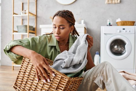 An African American woman with afro braids organizes laundry in a bathroom, sitting on the floor with a laundry basket.