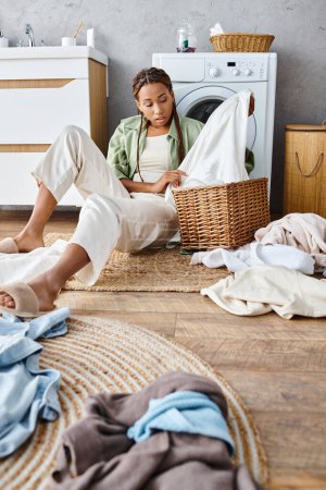 Foto de Una mujer afroamericana con trenzas afro se sienta en el suelo junto a una pila de ropa sucia, profundamente pensada en medio de la tarea del hogar. - Imagen libre de derechos