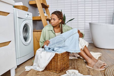 Eine afroamerikanische Frau mit Afro-Zöpfen sitzt auf dem Boden neben einem Wäschekorb im Badezimmer und sortiert Kleidung.