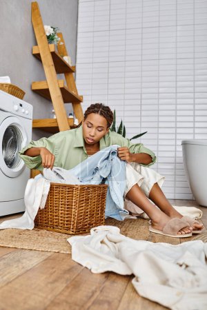 Una mujer afroamericana con trenzas afro sentada cerca de una lavadora, comprometida en la tarea de lavar la ropa en un baño.