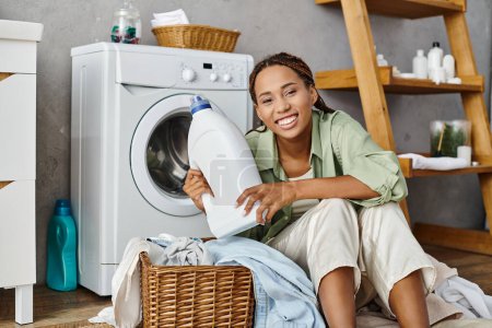 Una mujer afroamericana con trenzas afro se sienta al lado de una lavadora, lavando la ropa en un ambiente de baño.