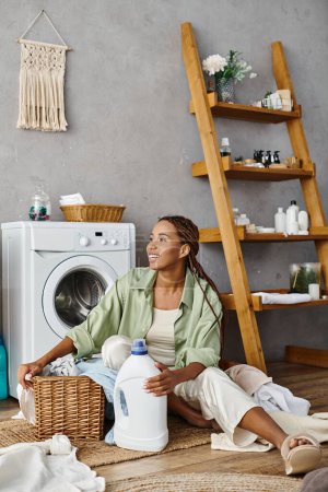 Foto de Una mujer afroamericana con trenzas afro se sienta tranquilamente en el suelo junto a una lavadora, lavando la ropa en un baño. - Imagen libre de derechos