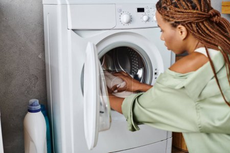 African American woman with afro braids putting clothes into a dryer in a bathroom while doing laundry.